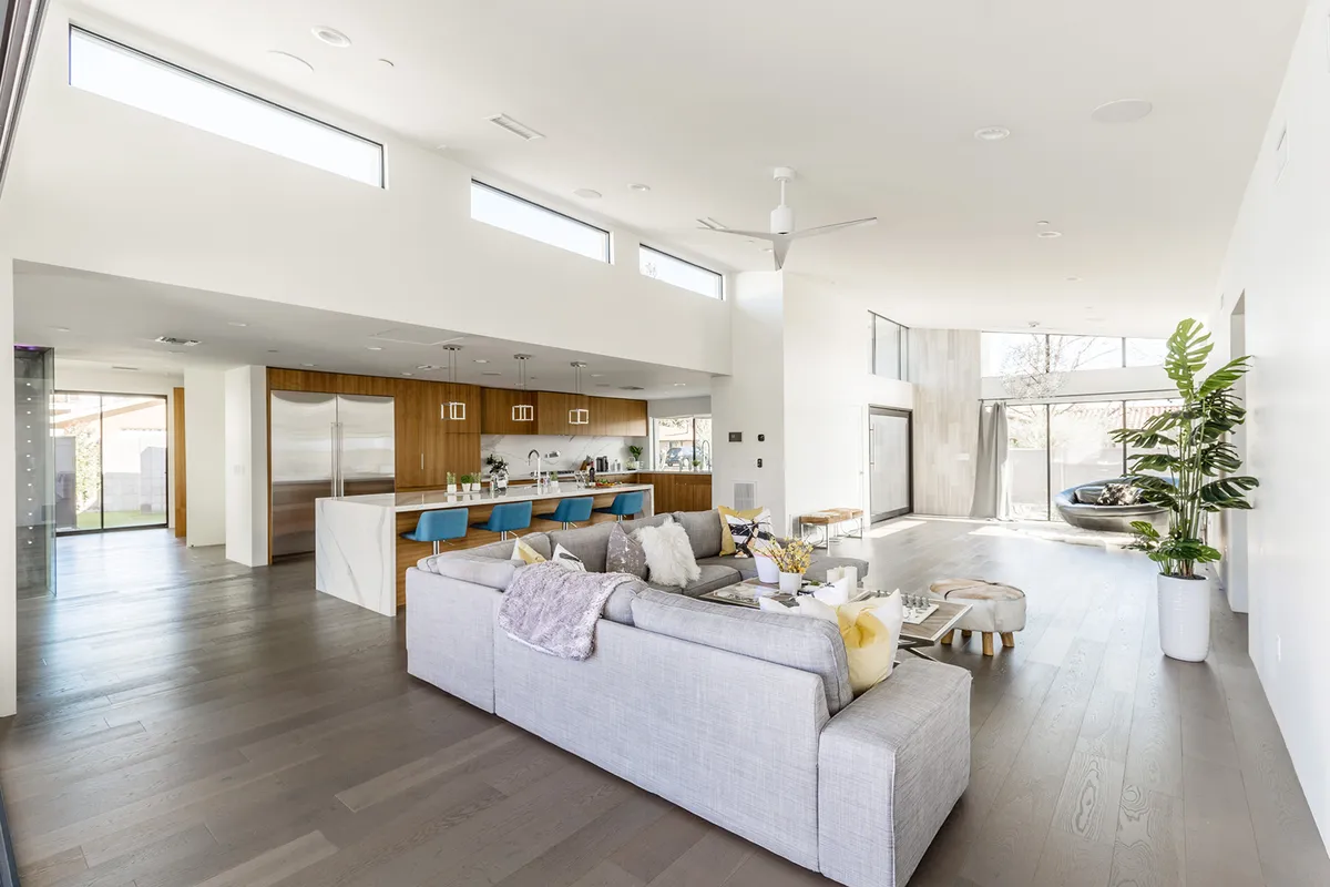 An inviting interior scene with a spacious living area, a modern kitchen featuring a sleek island, large windows letting in ample sunlight, and a comfortable sofa with cushions. The layout emphasizes both the kitchen and living room, with a clear view of the transition between spaces.