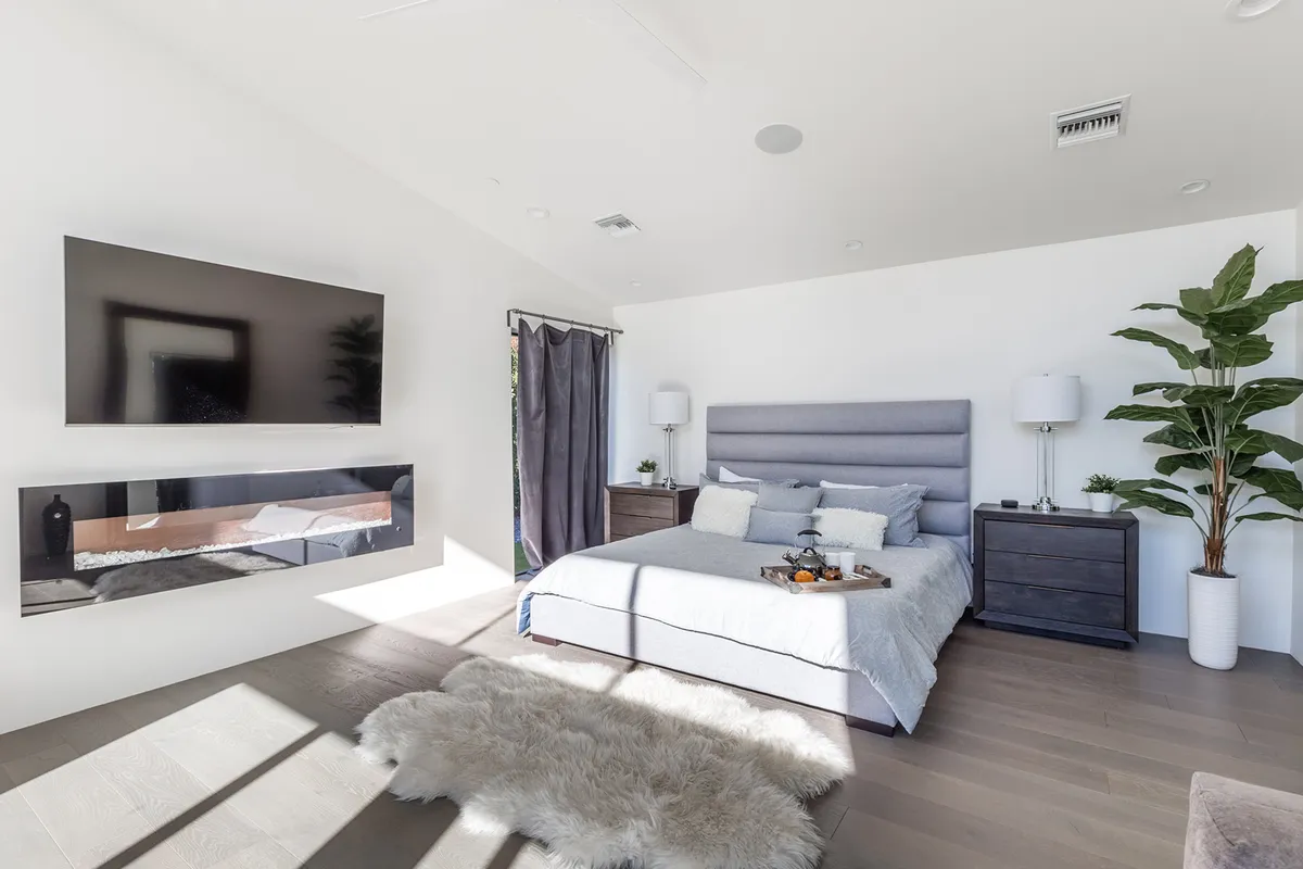 This image showcases a tranquil bedroom with a large bed, wall-mounted television, decorative plant, and nightstands, all arranged atop a textured rug. The soft lighting and neutral tones create a peaceful and inviting atmosphere, perfect for unwinding.