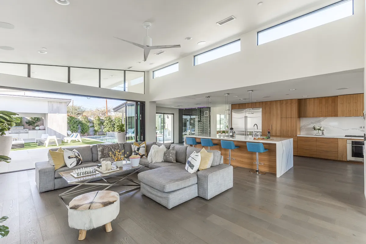 A spacious, open-concept living and kitchen area featuring a large sectional sofa, a prominent kitchen island with bar stools, and expansive glass windows that flood the room with natural light. The scene captures the seamless integration of the living, dining, and kitchen spaces, enhanced by the view of the outdoors just beyond.