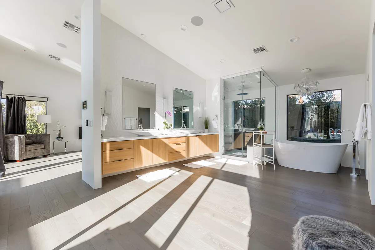 A modern bathroom with a double vanity—each with its own mirror—freestanding tub, and a glass shower enclosure. Large windows allow natural light to brighten the space, enhancing the serene and sophisticated mood. The bathroom is designed to accommodate multiple guests efficiently.