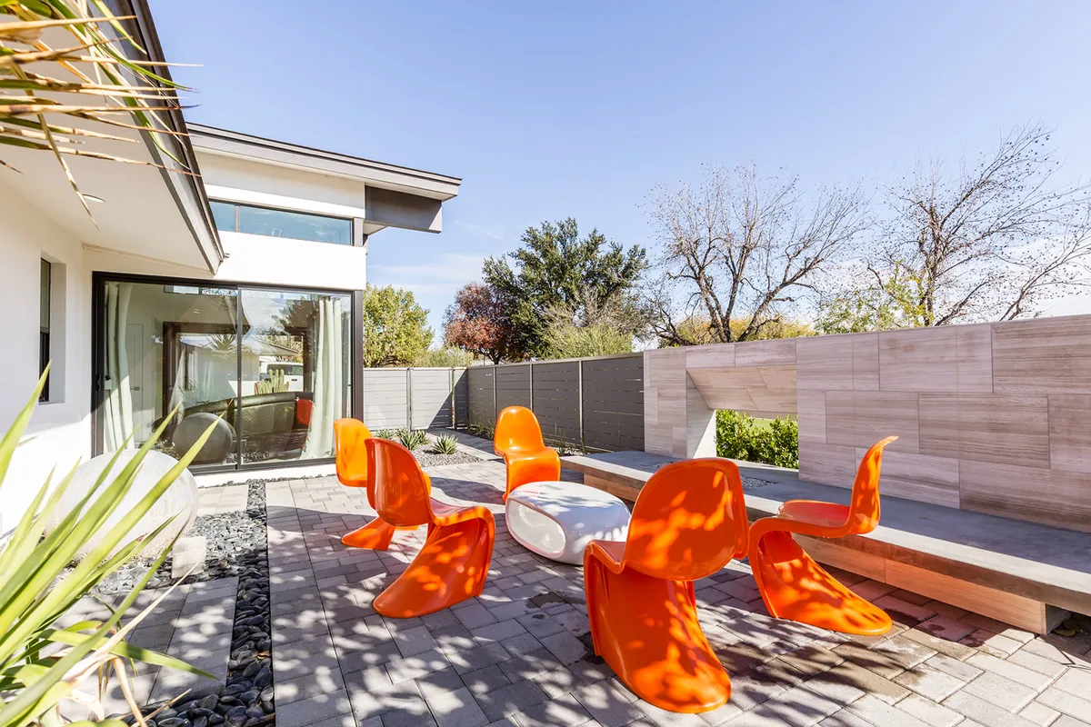 A vibrant outdoor dining area featuring bright orange chairs around a white circular table, set against modern architectural lines and lush landscaped greenery. The space is designed for lively social gatherings and sunset meals, offering a playful, festive atmosphere.