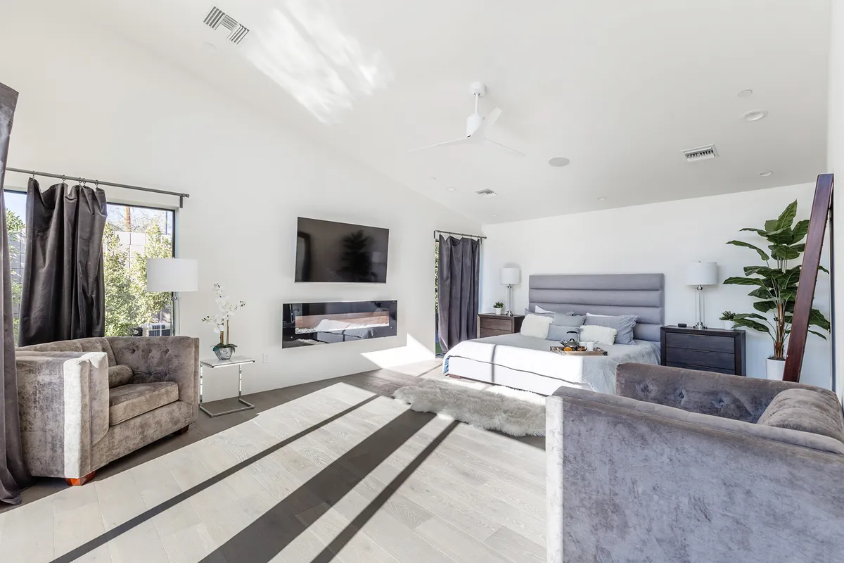 A calm and inviting bedroom featuring a comfortable bed, armchairs for lounging, a TV, and ample windows letting in natural light. Touches of greenery add freshness, and the arrangement encourages relaxation and personal space.