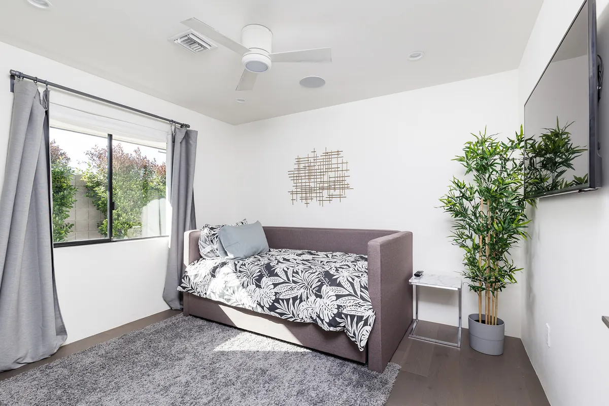 A cozy guest suite or secondary bedroom with a daybed adorned in floral bedding, a large window overlooking greenery, an indoor plant, and a wall-mounted TV. The setup is inviting for both adults and children, offering quiet space for relaxation, reading, or movie time.